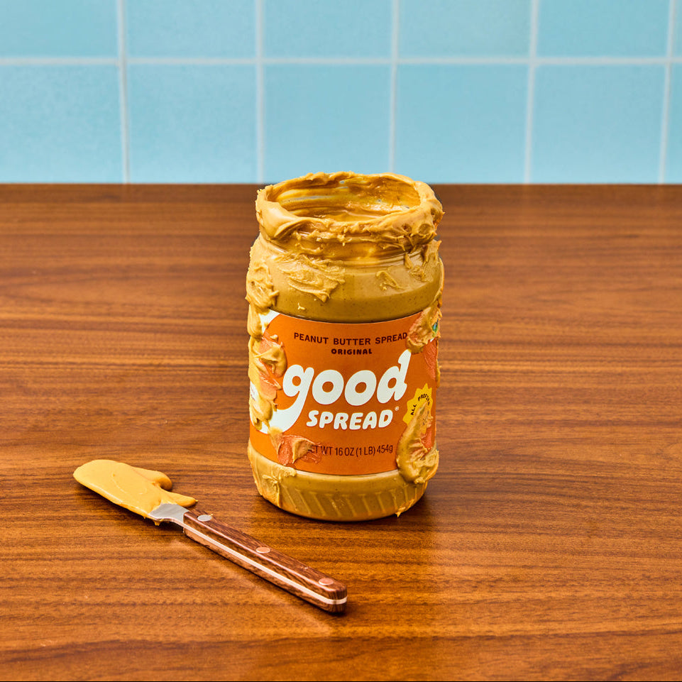 A jar of Good Spread peanut butter sits on a wooden tabletop with a matching spreader knife, set against a light blue tiled wall background.