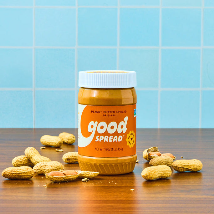 A jar of Good Spread peanut butter sits on a wooden table with scattered peanuts in front of a light blue tiled background.