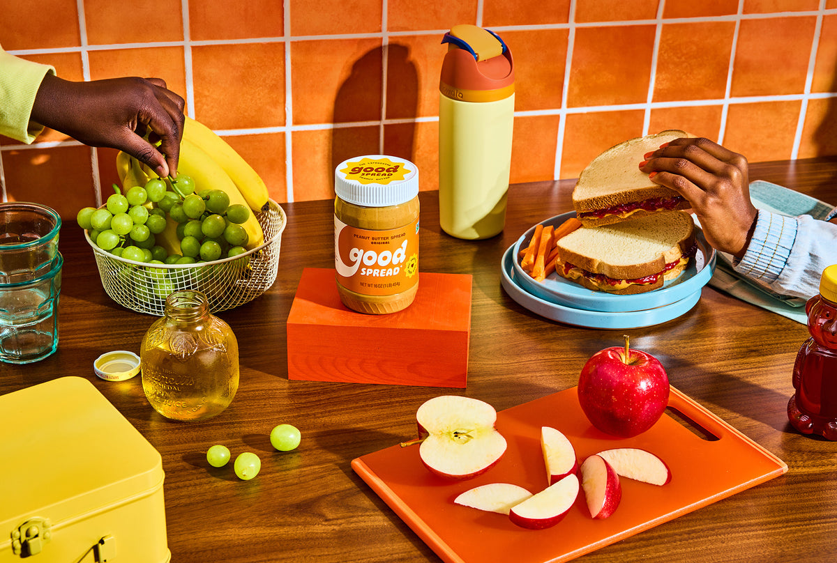 A styled lunch scene with a jar of Good Spread Peanut Butter centered on an orange block, surrounded by hands preparing a sandwich, eating green grapes, slicing an apple, and holding a yellow lunchbox against a retro orange tiled kitchen background.