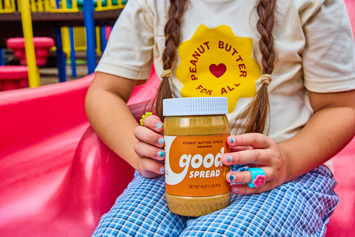 A child in a beige t-shirt and blue checkered pants holds a jar of Good Spread Peanut Butter in a colorful playground setting.