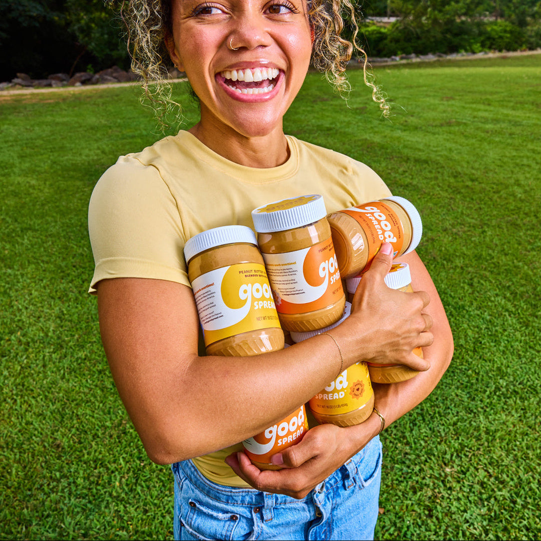 A smiling woman outdoors on a green lawn holds three jars of Good Spread Peanut Butter against her chest.