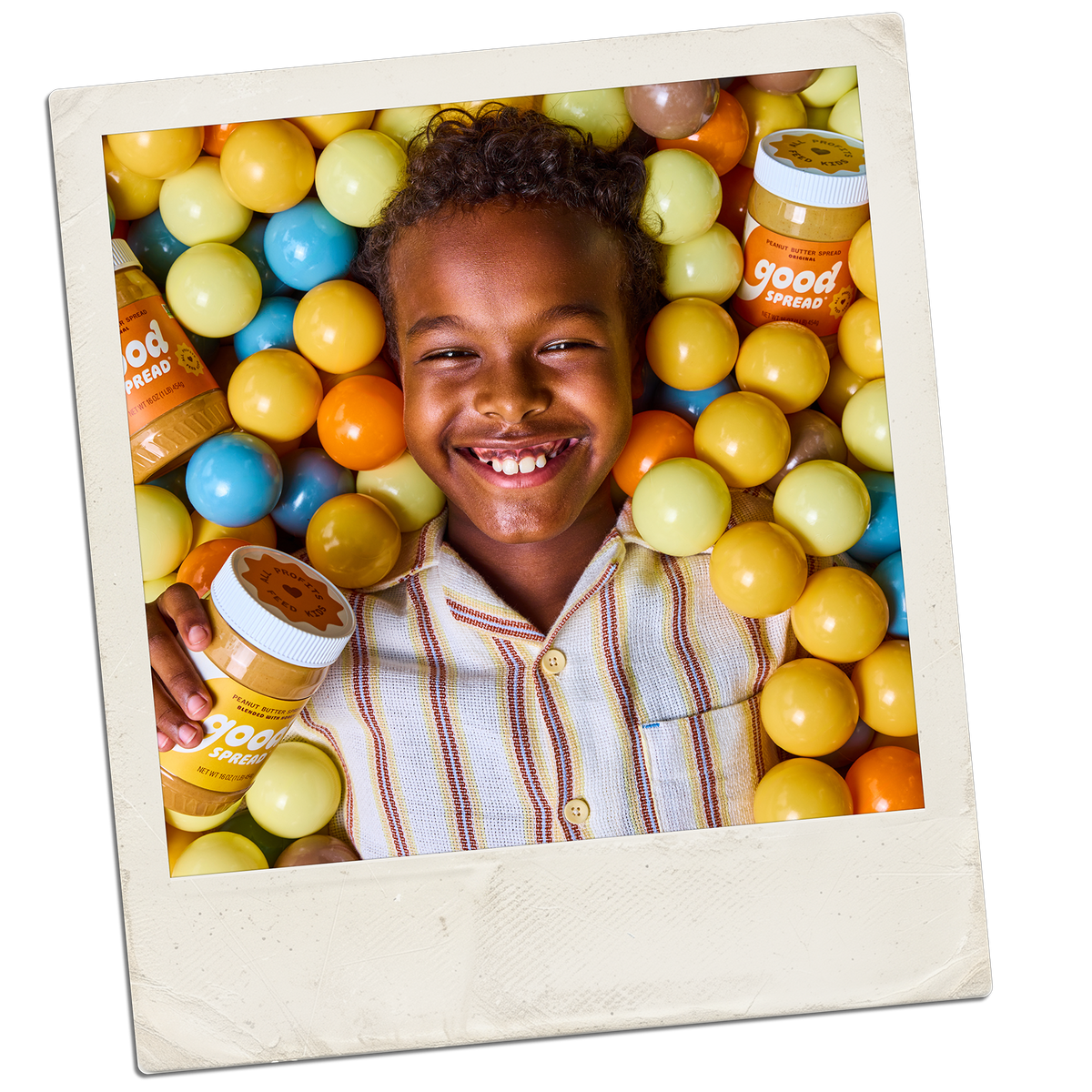 A smiling young boy lays in a ball pit filled with yellow, blue, and orange balls, holding a jar of Good Spread Peanut Butter. The photo is styled like a retro polaroid print with a cream border, set against a plain white background.